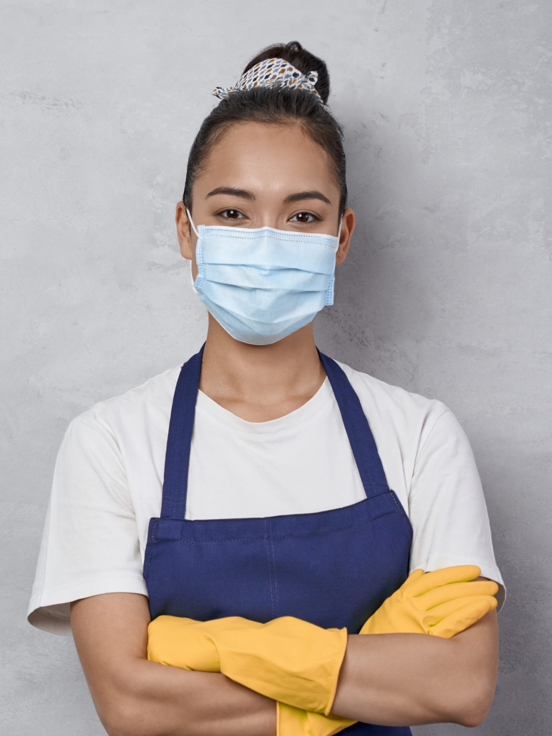 Confident cleaning lady wearing rubber gloves and medical protective face mask keeping arms crossed, looking at camera while standing against grey wall. Cleaning services during covid 19 pandemic